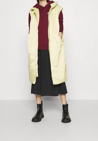 Ljusgul vadderad väst med huva, framknäppning, sidofickor, buren över en vinröd huvtröja och svart kjol, kombinerad med svarta ankelstövlar.