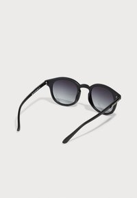 Schwarze Sonnenbrille mit abgerundetem Design, Verlaufsgläsern und dünnen schwarzen Bügeln. Ein Hardware-Detail am Bügel schafft einen Kontrast.