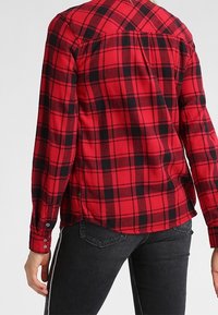 Camisa de franela a cuadros en rojo y negro, con mangas largas y puños abotonados, que presenta un diseño de yugo en los hombros y aberturas en los laterales del dobladillo.