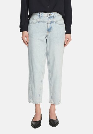 Kobieta ubrana w jasnoniebieskie, zwężane jeansy z wysokim stanem, czarną bluzkę z długim rękawem i czarne czółenka typu slingback z ostro zakończonym noskiem, stojąca.