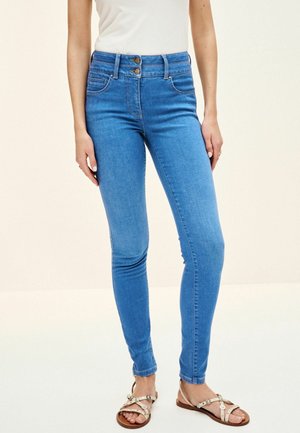 Donna che indossa jeans skinny a vita alta azzurri con chiusura a doppio bottone e sandali beige con borchie decorative.