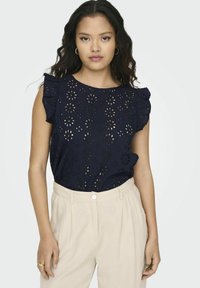 Top blu navy con motivo floreale a taglio, scollatura rotonda e maniche corte a volant. Abbinato a pantaloni beige chiaro con chiusura frontale a bottone.