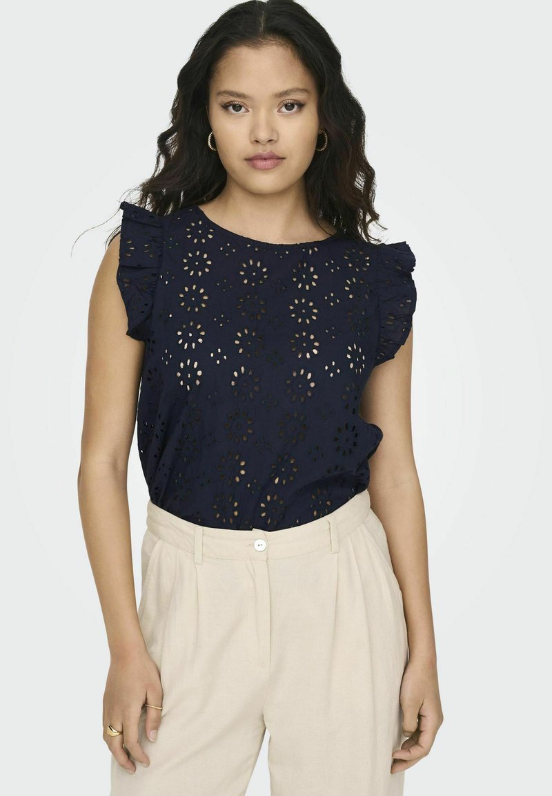 Top blu navy con motivo floreale a taglio, scollatura rotonda e maniche corte a volant. Abbinato a pantaloni beige chiaro con chiusura frontale a bottone.