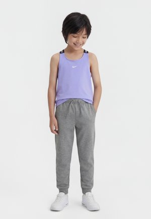 Barn med kort hår iført en lavendel tanktop og grå joggingbukser, står med hænderne i lommerne og smilende nedad.