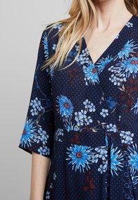 Mörkblå klänning med blommönster och prickar. V-ringning, korta ärmar, markerad midja och klara blå blommönster. Mjuk tygkvalitet.