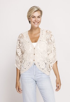Smiling woman wearing a beige crochet buttoned cardigan over a white top and light blue jeans, standing against a plain background.