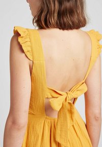 Robe jaune avec des bretelles volantes, design dos nu et détail de nœud noué. Le tissu a une apparence texturée et froissée.