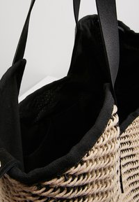 Sac cabas tissé beige et noir avec doublure en tissu noir et poignées en cuir noir suspendu devant un fond uni.