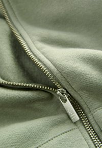 Grüne Sweatshirtstoff mit einem silbernen Reißverschluss, an dem ein Etikett mit dem Aufdruck "self" befestigt ist. Nahaufnahme von Textur und Nähten.