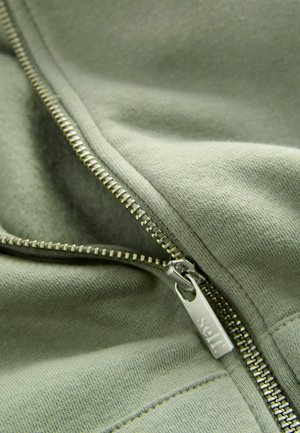 Groene sweatshirtstof met een zilveren ritssluiting, voorzien van een label met de tekst "self" eraan bevestigd aan de rits. Close-up van de textuur en stiksels.