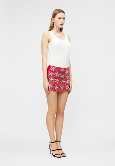 Γυναίκα με μακριά ίσια μαλλιά, ντυμένη με λευκό αμάνικο τοπ, κόκκινη μίνι φούστα με σχέδια από μπλε αστερίες και μαύρα σανδάλια με τακούνι.