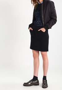Une veste bomber matelassée noire sur un pull à pois bleu marine, associée à une jupe sombre et des chaussures noires brillantes, avec une semelle épaisse et une bride.