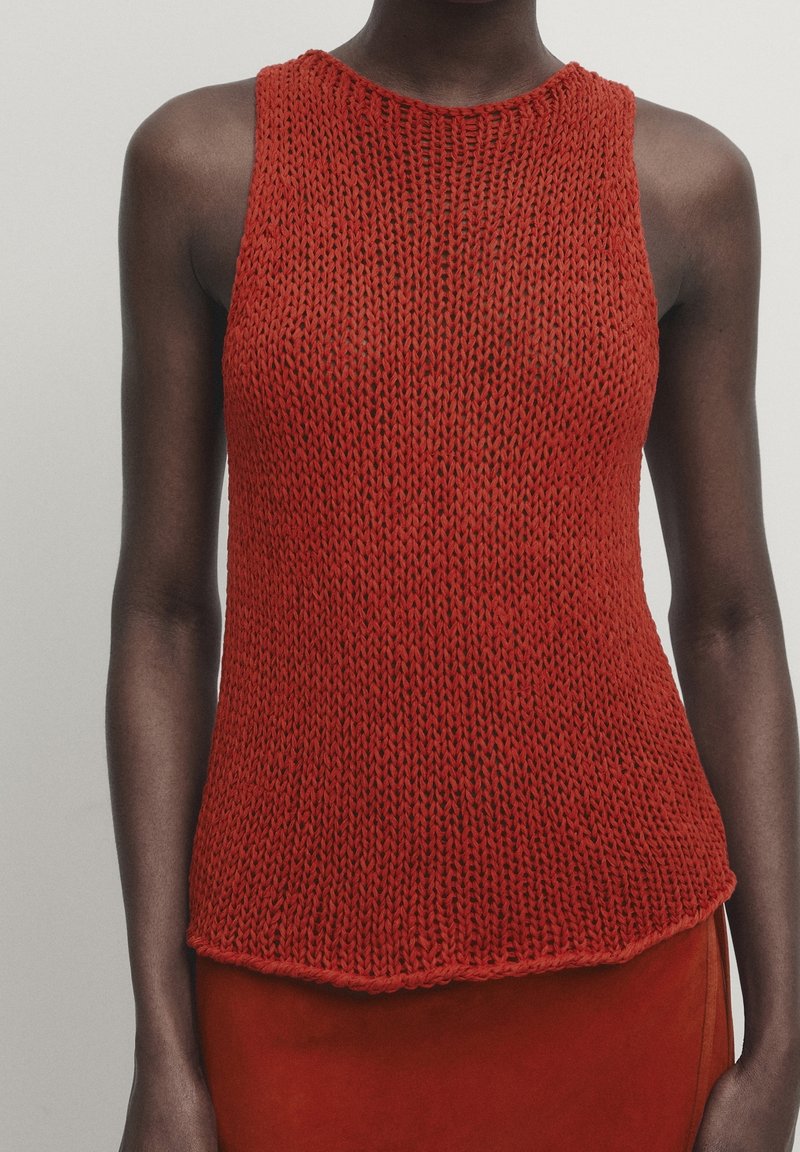 Punainen neule ilman hihoja, jossa on pyöreä pääntie. Ominaisuuksiin kuuluu löysä kudontatekstuuri ja hieman kaareva helma. Materiaali näyttää pehmeältä ja hengittävältä.