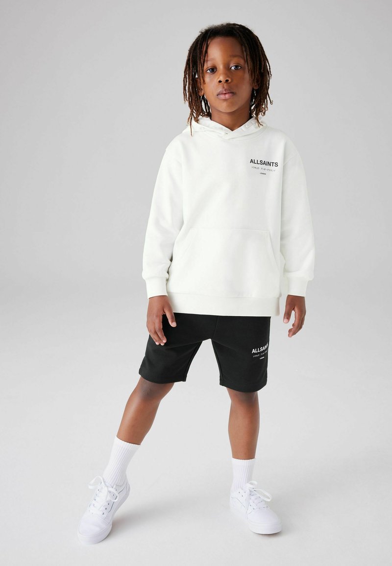Vit hoodie med framficka och svart textlogga, i kombination med svarta shorts som har samma logga. Vita sneakers och sockar.
