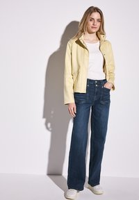 Chaqueta de cuero amarillo claro con botones a presión, llevada sobre una camisa blanca; combinada con jeans de pierna ancha de color azul oscuro y zapatillas blancas.