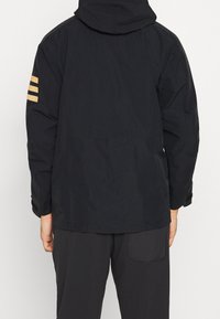 Veste noire avec capuche, ornée de trois rayures dorées horizontales sur la manche gauche et d'un tissu lisse et texturé.