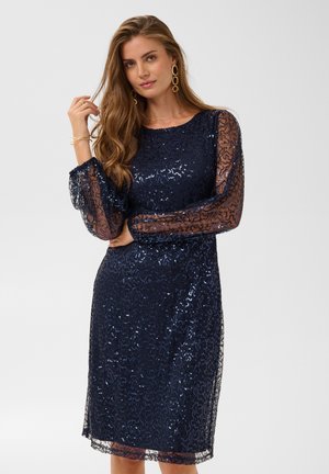 Femme portant une robe bleu marine à sequins avec des manches longues transparentes, debout, une main touchant ses cheveux et l'autre reposant sur sa taille.