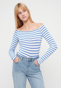 Μπλε και λευκή ριγέ μπλούζα με ανοιχτούς ώμους και μακριά μανίκια, κατασκευασμένη από απαλό ύφασμα, σε συνδυασμό με ψηλόμεσο ανοιχτό μπλε τζιν παντελόνι.