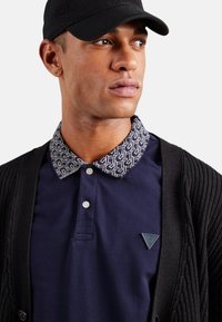 Polo azul marino con collar estampado, cárdigan negro y acento de logotipo triangular. El collar presenta un diseño geométrico en blanco y azul.