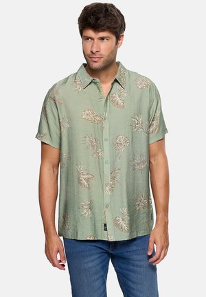 Hombre vistiendo una camisa de manga corta verde con un patrón de hojas tropicales beige y jeans azules, de pie contra un fondo liso.