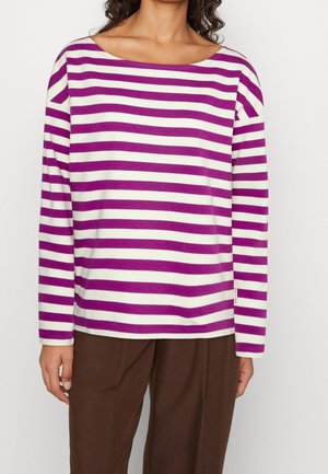 Femme portant un haut à manches longues blanc avec des rayures horizontales violettes, associé à un pantalon marron, sur un fond uni.