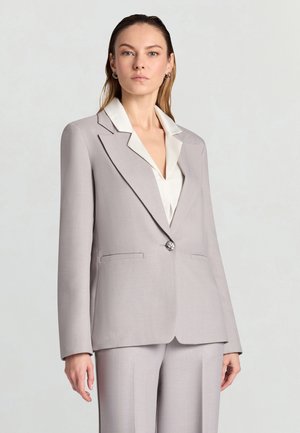 Donna che indossa un completo su misura grigio chiaro con camicia a collo bianco, in piedi davanti a uno sfondo chiaro e semplice, rivolta verso l'obiettivo.