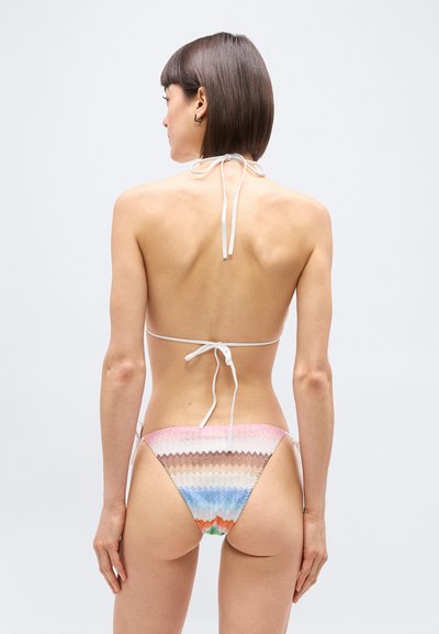 Γυναίκα με κοντά καστανά μαλλιά που φοράει μαγιό με πολύχρωμο μοτίβο ζιγκ-ζαγκ στο κάτω μέρος και λευκό μπικίνι κορδόνι πάνω μέρος, εμφανίζεται από πίσω.