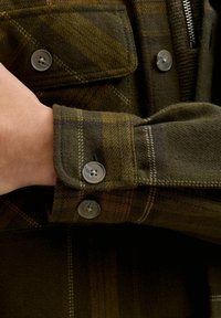Grün-braun karierte Hemd aus einem weichen, strukturierten Stoff mit Metallknöpfen und Knopfmanschetten-Details.