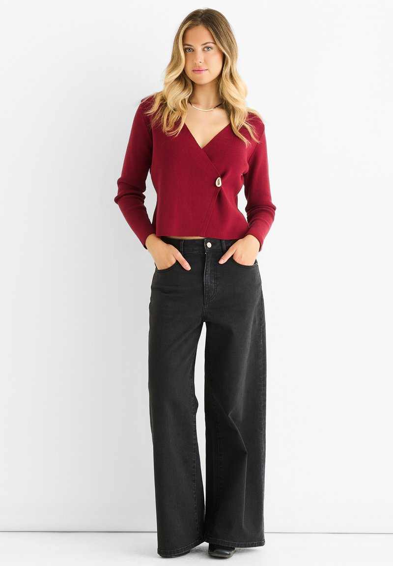 Maglione avvolgente bordeaux con un bottone decorativo, abbinato a jeans neri a vita alta e gamba larga. Texture liscia e design aderente.