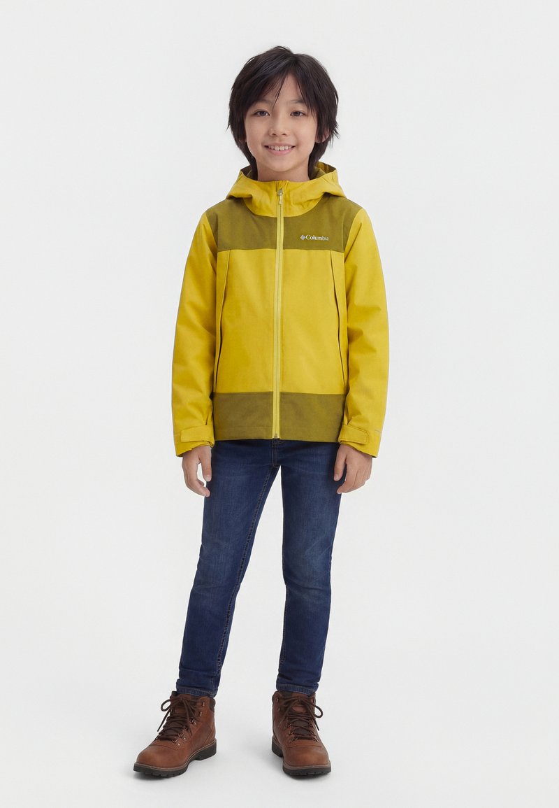 Αγόρι όρθιο και χαμογελαστό, φοράει κίτρινο και ελιάς μπουφάν με κουκούλα, μπλε τζιν και καφέ μπότες με κορδόνια, πάνω σε απλό φόντο.