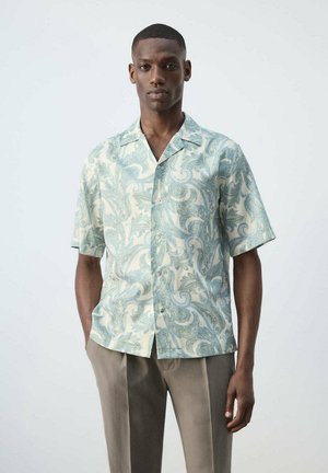 Joven de pie con una camisa de manga corta de botones estilo paisley en azul claro y beige y pantalones beige sobre un fondo liso.