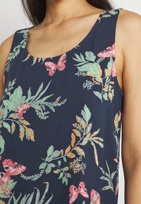 Top in tessuto navajo con fiori, presenta foglie verdi e farfalle rosa, con scollatura a barchetta e texture leggera.