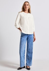 Weißes Hemd mit Raffung am Ausschnitt, kombiniert mit hellblauen weit geschnittenen Jeans. Das Model steht in schwarzen Ballerinas.