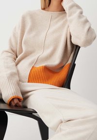 Beżowy sweter dzianinowy z prążkowanym pomarańczowym akcentem w talii, z długimi rękawami i okrągłym dekoltem, noszony na kremowych spodniach.