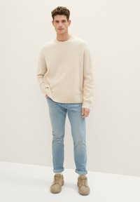 Pulover tricotat de un bej deschis, cu guler rotund, asortat cu blugi albaștri deschiși. Modelul poartă adidași din suede bej. Design simplu și potrivit.
