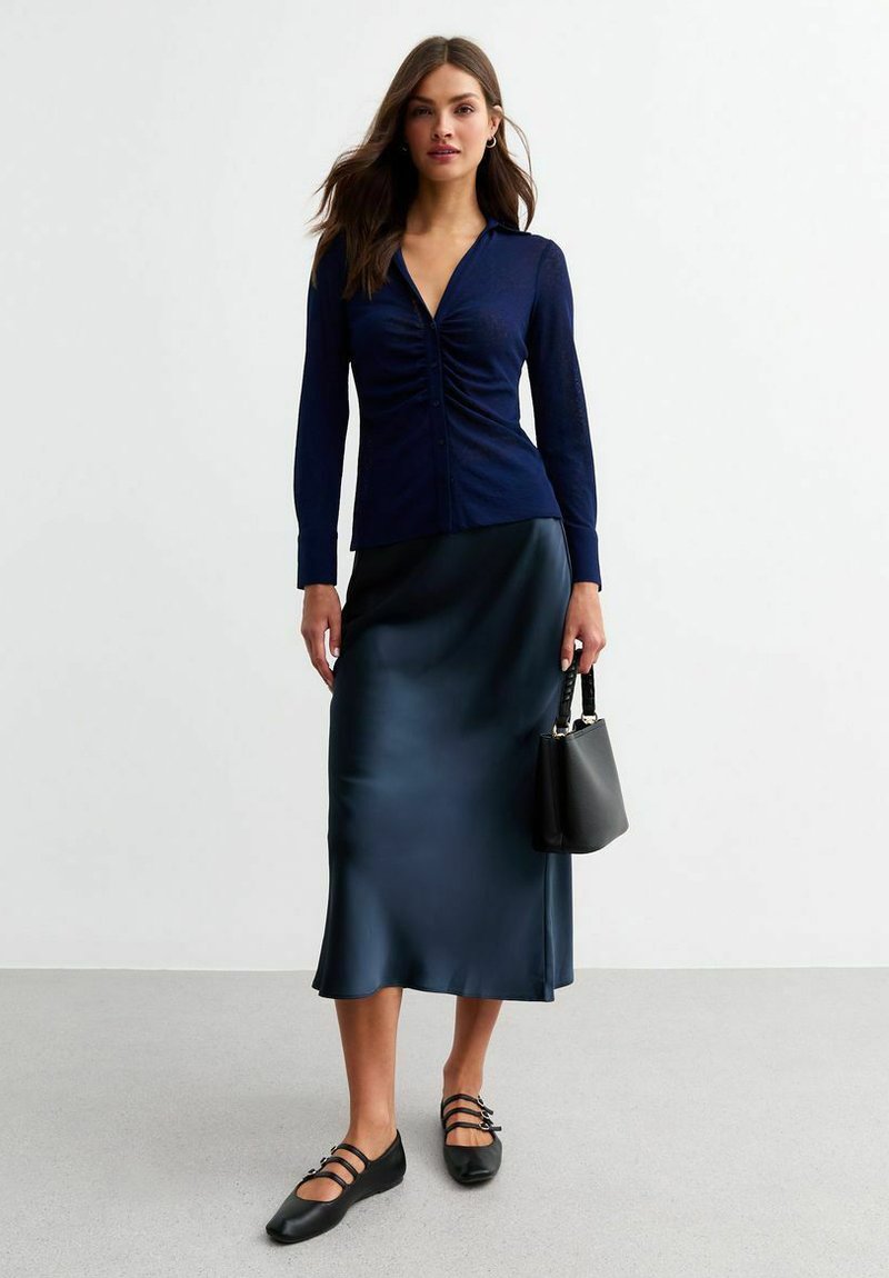Haut à manches longues bleu marine avec ruché à l'avant, associé à une jupe en satin arrivant au genou. Sac à main noir et ballerines avec des détails de sangle.