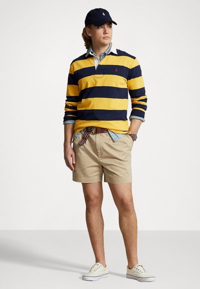 Polo s dugim rukavima u mornarsko plavoj i žutoj prugastoj kombinaciji, preko svijetloplavog šambray košulje, u paru s kaki šortsevima i bijelim tenisicama. Tamna kapa naglašava izgled.