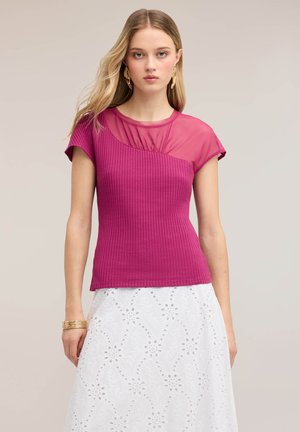 Kvinde med langt blond hår iført en kortærmet magenta ribstrikket top med gennemsigtig bærestykke og hvid øjletsblonde nederdel, stående mod en neutral baggrund.