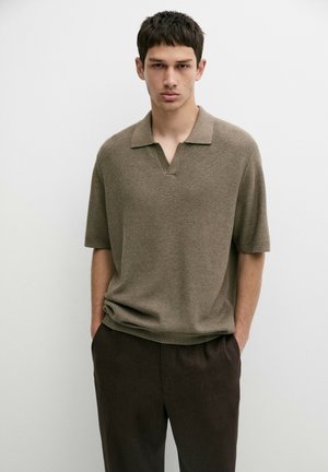 Jonge man draagt een kort mouw bruin polo met structuur en donkerbruine broek, staand met handen in de zakken tegen een effen witte achtergrond.