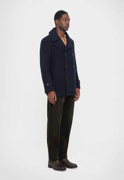 Peacoat de lã azul-marinho com um design de duplo peito, grandes lapelas e bolsos laterais. Combinado com calças de veludo cotelê castanhas e botas castanhas escuras.