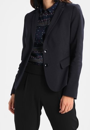 Mujer que lleva una blazer azul marino sobre una camisa azul marino con estampado y pantalones negros a medida, de pie con una mano relajada a su lado.