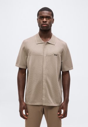 Νέος άνδρας που φοράει μπεζ κοντομάνικο πόλο με γιακά, μικρό λογότυπο "BOSS" στο στήθος, και ταιριαστό μπεζ παντελόνι, στέκεται μπροστά σε απλό φόντο.