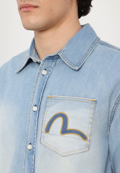 Camicia in denim azzurra chiara con colletto a bottoni, dotata di una tasca sul petto decorata con un motivo di cuciture curve blu e gialle.