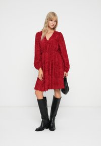 Rotes, strukturiertes Kleid mit V-Ausschnitt, langen Puffärmeln und gebundenem Taillengürtel. Getragen mit schwarzen kniehohen Stiefeln und einer kleinen schwarzen Tasche.