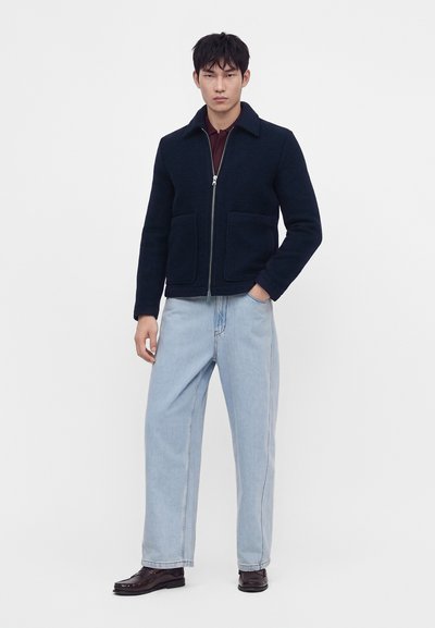 Jeune homme debout portant une veste zippée sombre, un jean bleu clair à jambes larges, des chaussures foncées, une main dans la poche, sur un fond uni.