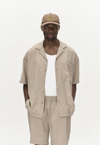 Camicia a maniche corte beige chiaro con bottoni e taschino, abbinata a pantaloni corti coordinati. Un cappello beige presenta la scritta "Eagle Creek Wilderness" in giallo.