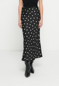 Svart maxi skjørt med polkadotter, glatt tekstur og utsvingt kant. Brukt med tettsittende svart topp og knelange svarte støvler.