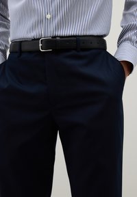 Marineblaue Hose mit Seitentaschen, kombiniert mit einem schwarzen Leder-Gürtel mit einer silbernen Schnalle. Ein blau-weiß gestreiftes Hemd mit Knöpfen ist sichtbar.