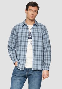 Blauw geruite button-up shirt met donkere lijnen, lange mouwen en een borstzak. Ggedragen over een wit graphic t-shirt en gecombineerd met blauwe jeans.