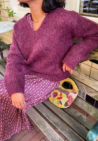 Pull en tricot bordeaux avec un col en V, manches longues. Jupe violette à motifs, sac à main décoratif jaune avec perles et accents. Banc en bois.